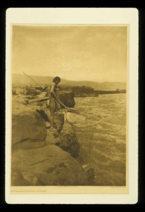 Sepia toned image of a person holding a large net standing next to a body of water. 