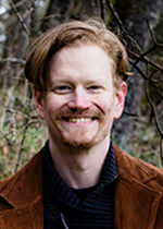 Profile picture of John Taylor with medium length brown reddish hair with a facial beard. Wearing a black knitted shirt and a burnt umber jacket.