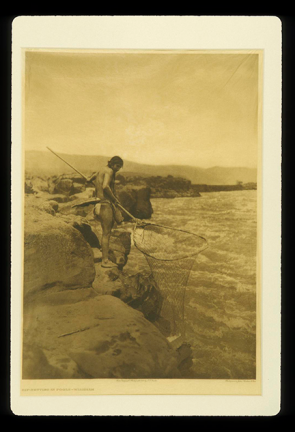 A person stands on a flat, rocky coastline, holding a large net over the water. 