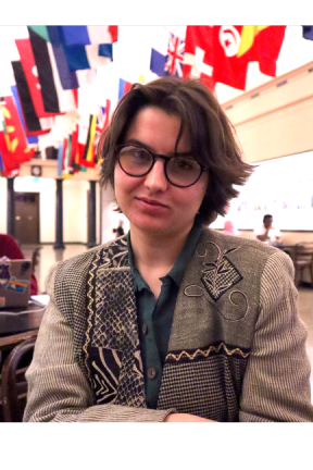 A non-binary person with short dark hair, light skin tone and black-rimmed glasses wits under a display of national flags. 