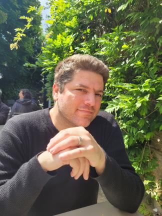 A person with light skin tone, a short beard and short, light brown hair sits in front of a large, leafy wall.