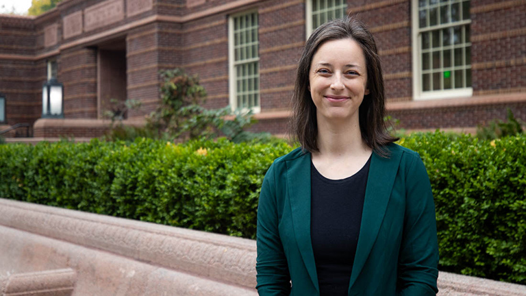 A woman with shoulder-length dark hair and light skin tone stands smiling in front of a library. 