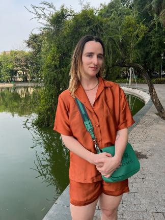 A person with shoulder-length blonde hair and light skin tone stands wearing an orange jumper and green bag. 