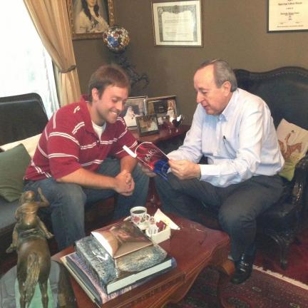 Two men sit in a living room and look at a book. 