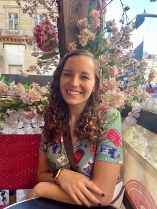 A female-presenting person with long, curly blonde hair and light skin tone smiles , seated in front of a bunch of flower blossoms.