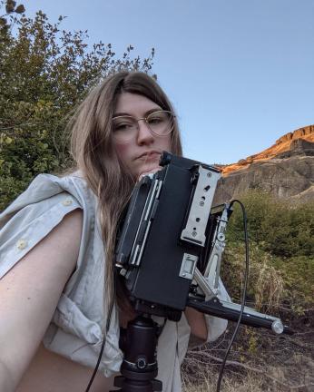 Color photo of a non-binary person with light skin tone and long brown hair. They are outside in a desert-like space, and hold a large film camera.