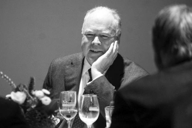 Black and white photo of a man seated at a table. He rests his head on his left hand and is in conversation with a person who has their back to the camera. 