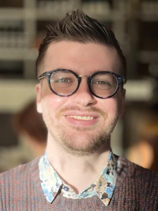 A person with dark hair, a beard, light skin tone and glasses smiles into the camera.