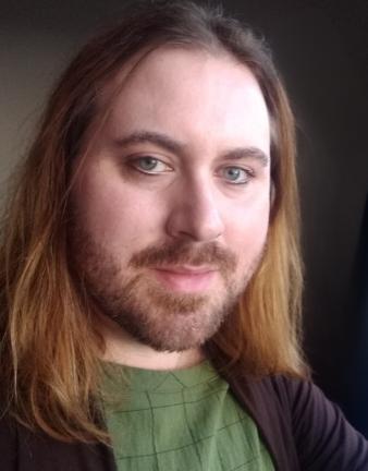 Color photo of a non-binary person with light skin tone and shoulder-length brown hair. They have a mustache and wear a green tee-shirt. 