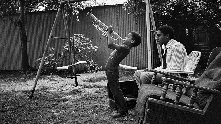 a boy blowing a trumpet in a yard with a swing set and older man sitting in a lawn chair