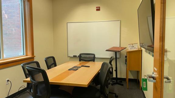 View into a study room with a conference table and chairs in front of a wall mounted screen and whiteboard.