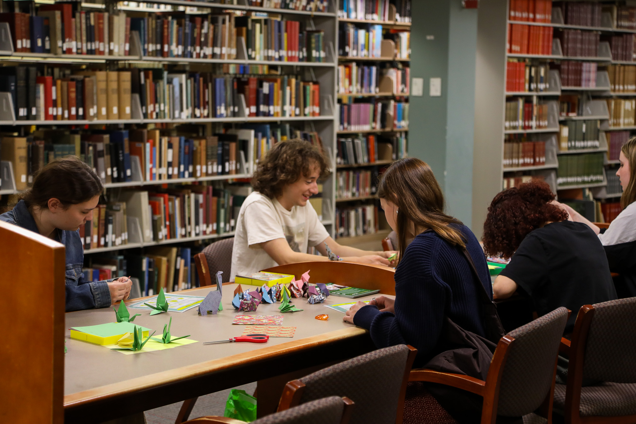 Students folding craft paper into origami ducks. Library bookshelves are visible in the background.