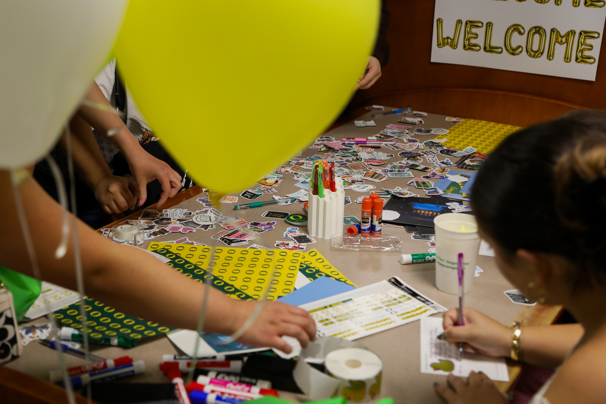 Close-up of stickers, pens, and glue-sticks on a crafting station table; there are balloons in the foreground.