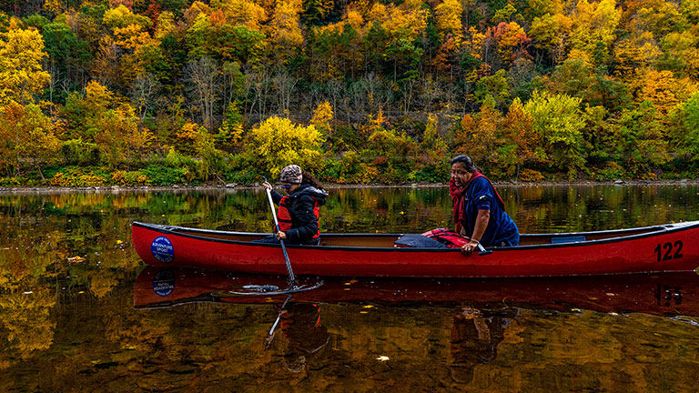 Photo of two people in a canoe on a body of water with trees in full fall foliage behind them.