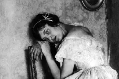 A young woman wears a white gown and rests her head on the back of a wooden chair. 