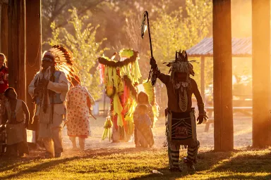 A person dressed in regalia holds a staff high above their head, while golden sunlight streams in from the right side of the image. Children and other adults follow. 