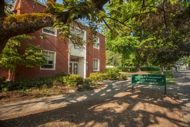 A square, two-story brick building has white double doors and sits among large trees. A green sign reads MarAbel B. Forhnmayer Music Building Beall Concert Hall.