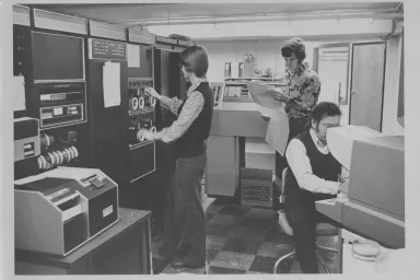 Three people wear 1970s clothes and work on a giant computer.