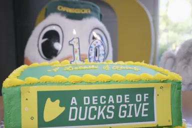 Oregon Duck standing beside a celebratory cake with "10" candles and frosting that spells out, "A Decade of Ducks Give."