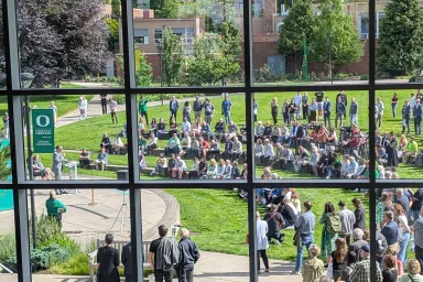 View through an upper-story window grid at many people gathered on a campus lawn, listening to Karl Scholz speak.
