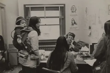 Illustrations of Native American men are displayed on the wall behind a group of women gathered around an office desk.