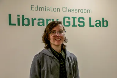Person is in jean jacket standing in front of signage for the library GIS lab.