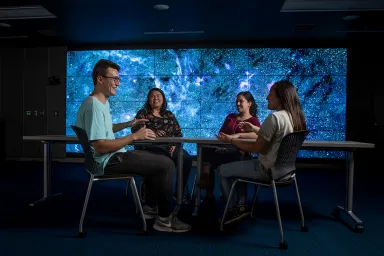 Four students sitting at a Science Library table with a blue image of the Milky Way on a digital display screen in the background.
