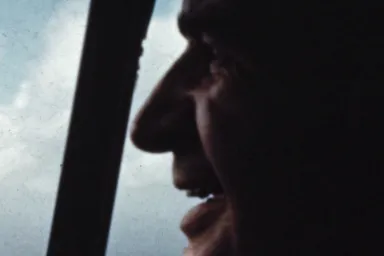 Man's face in profile next to an aircraft's windshield with blue sky and clouds visible outside.