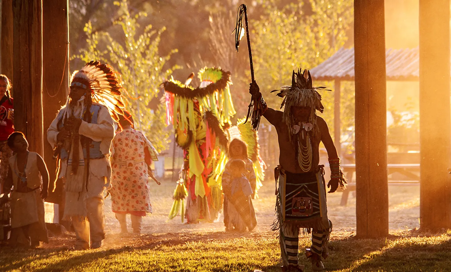 A person dressed in regalia holds a staff high above their head, while golden sunlight streams in from the right side of the image. Children and other adults follow. 