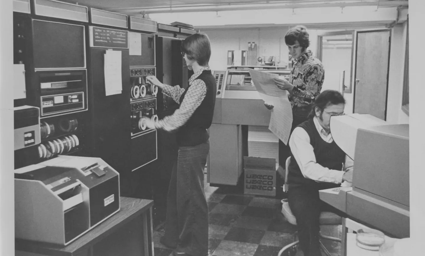 Three people wear 1970s clothes and work on a giant computer.