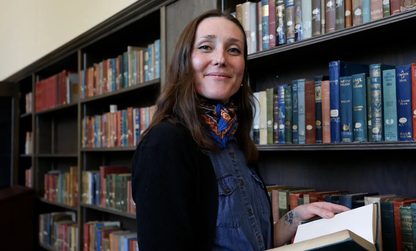 Emily Moore holding an open book, standing next to a bookcase in Special Collections.