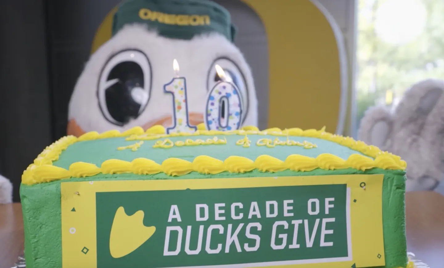 Oregon Duck standing beside a celebratory cake with "10" candles and frosting that spells out, "A Decade of Ducks Give."