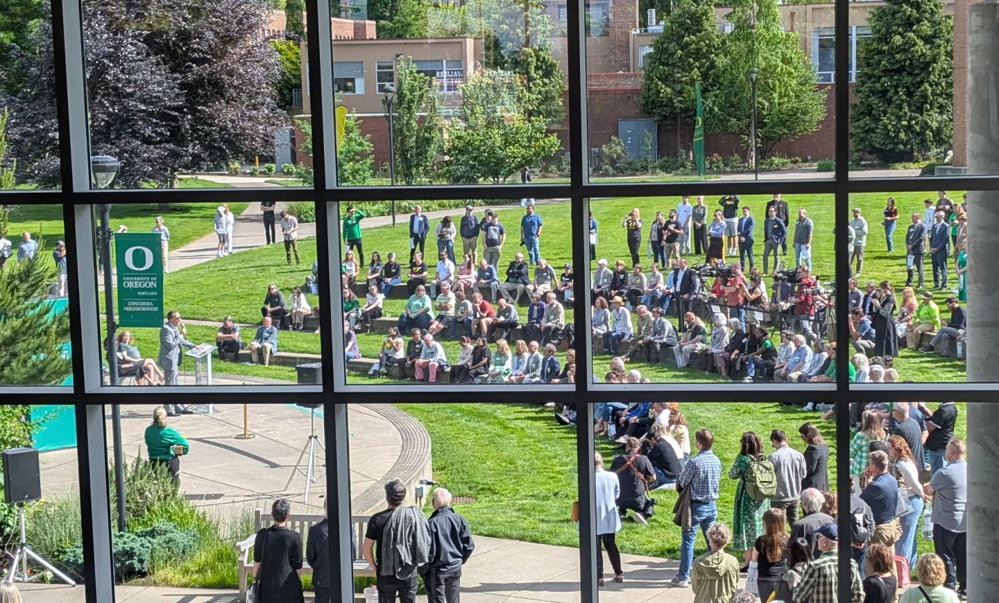 View through an upper-story window grid at many people gathered on a campus lawn, listening to Karl Scholz speak.
