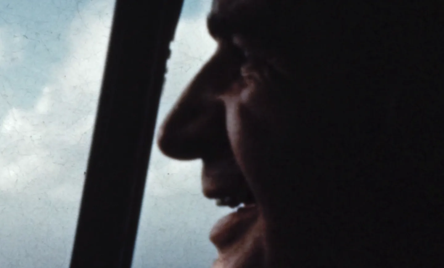 Man's face in profile next to an aircraft's windshield with blue sky and clouds visible outside.