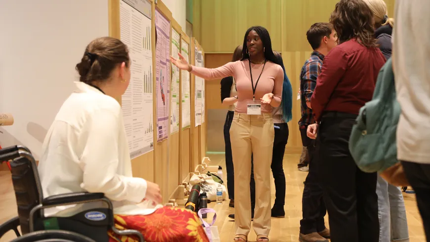 A Black woman presenting her research poster to five symposium attendees.