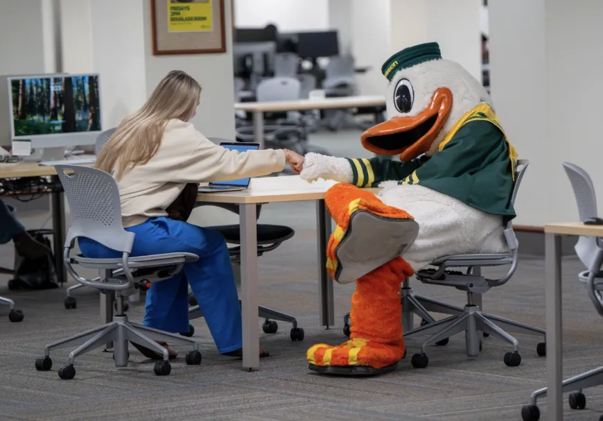 The Duck mascot fist-bumps a student studying at a Knight Library table.
