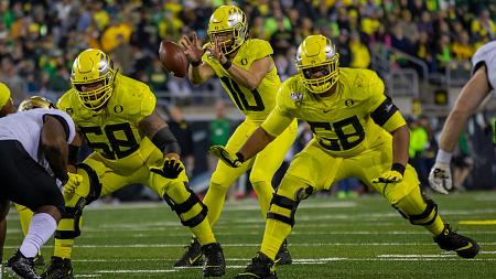 QB Justin Herbert receiving a snap as linemen Shane Lemieux and Penei Sewell provide protection during a football game.