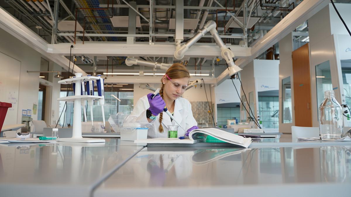 Woman wearing safety goggles and lab coat pipetting green solution from a beaker in a lab at the Knight Campus.