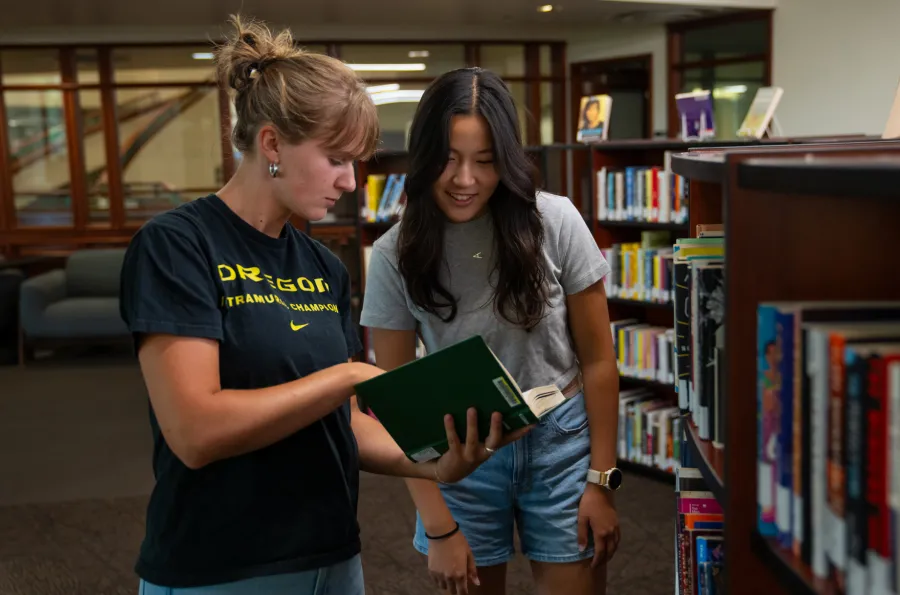 Two students looking at a book in front of a bookshelf