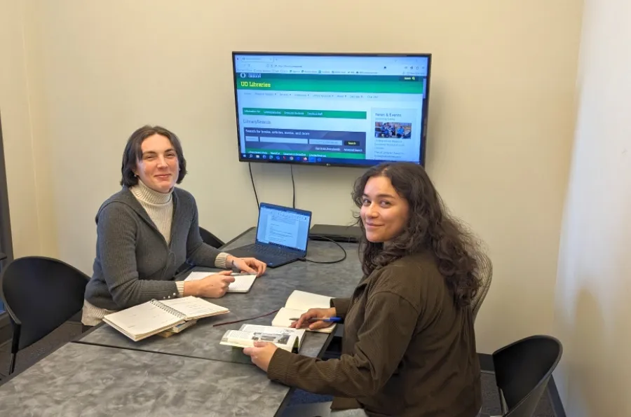 Two students in a study room with the library website up on the TV that is connected to a laptop